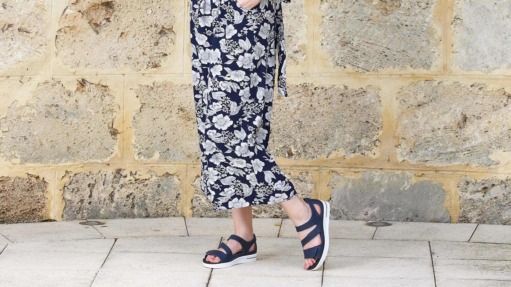Lady walking in front of limestone background wearing navy Klouds Patience sandals